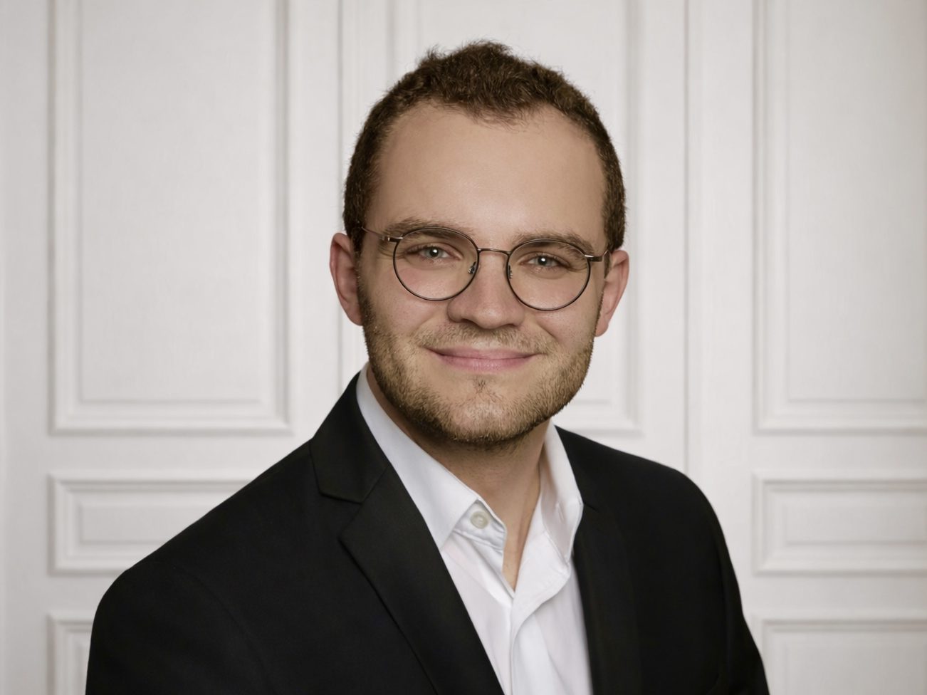 OCULUS-Immobilien Inhaber Bernhard Paulus Ein junger Mann mit Brille, lächelnd in formeller Kleidung vor einer neutralen Wand.