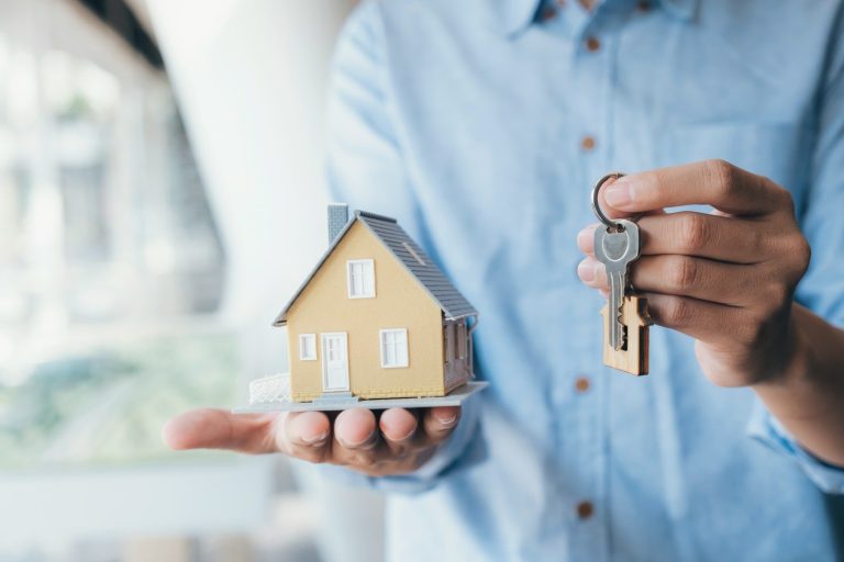 Symbolbild einer werthaltigen Immobilie Eine Person hält ein kleines gelbes Hausmodell und Schlüssel in der Hand.