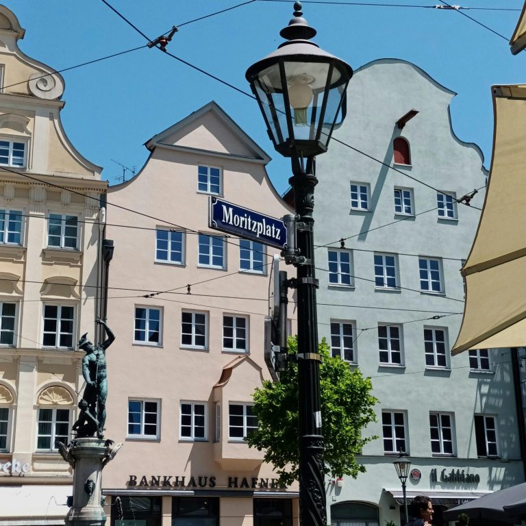 Augsburger Impressionen Farbenfrohe Gebäude an einem Platz mit Straßenlaterne und grüner Pflanze im Vordergrund.